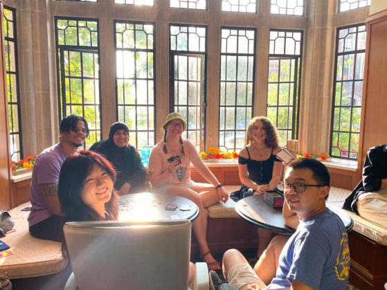 a group of six students sit smiling together in front of a large window