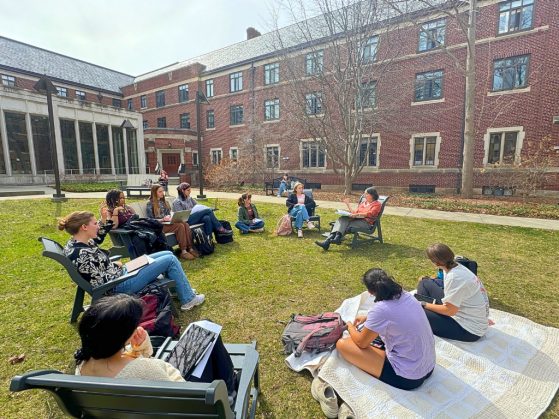 a class sits together outside, they are seated in chairs or on the ground on blankets