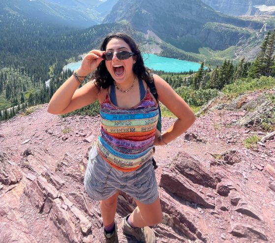 female in rainbow tie-dye top on a mountain smiling at camera