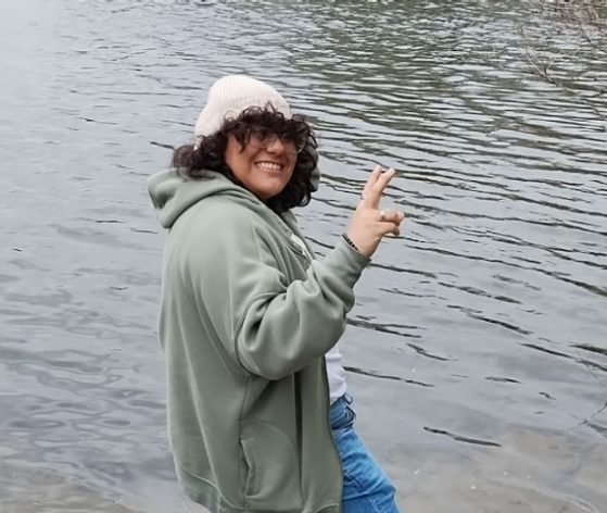 female in green jacket and jeans at a river smiling at camera