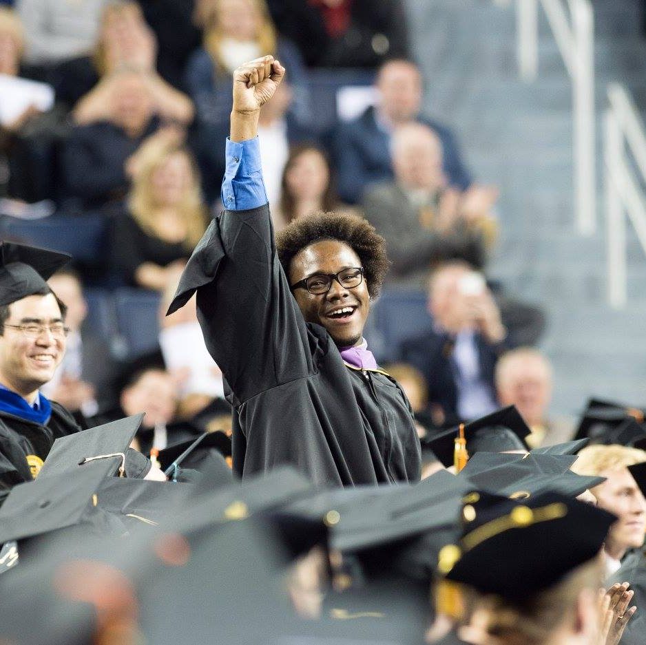 One male black graduate stands  in focus among blurred sitting graduates, smiling with his fist punching the air in celebration