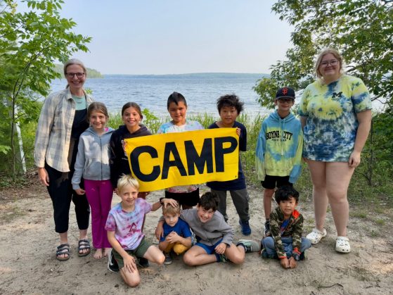 Children holding a sign that reads "Camp" along a lake