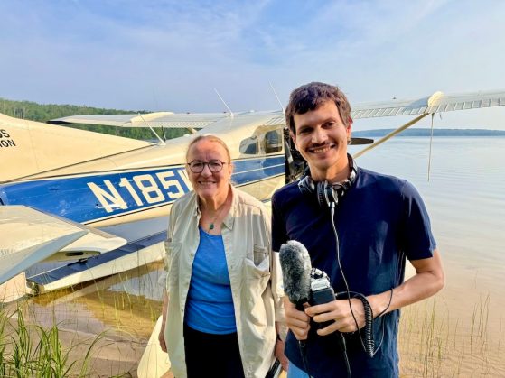 Two people standing along a lake shore with a seaplane in the background