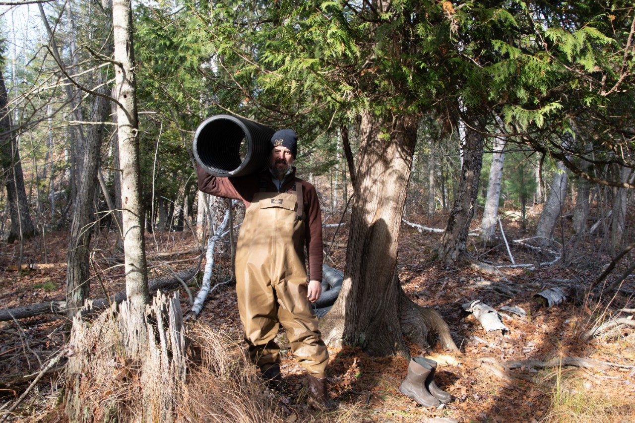 Man wearing waders in the woods and holding a large plastic pipe over his shoulder
