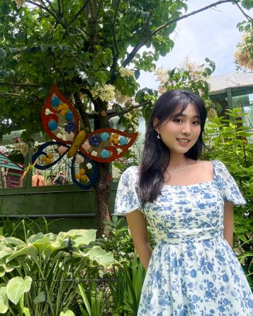 A woman in a floral dress stands by a lush garden with a vibrant, decorative butterfly on a tree. The scene is bright and cheerful.