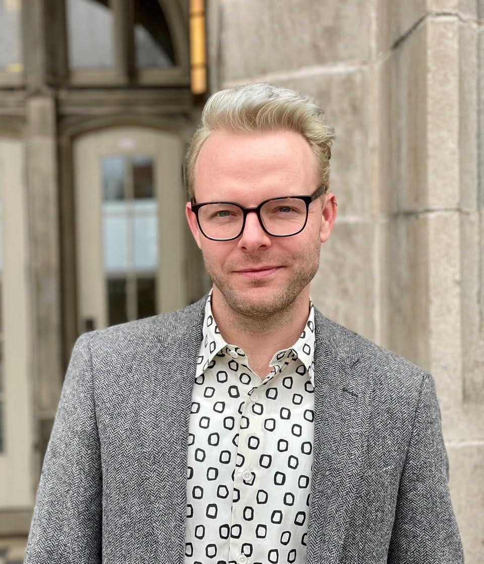 Professor Grzanka smiles in a black and white shirt and gray jacket against an outdoor backgroung