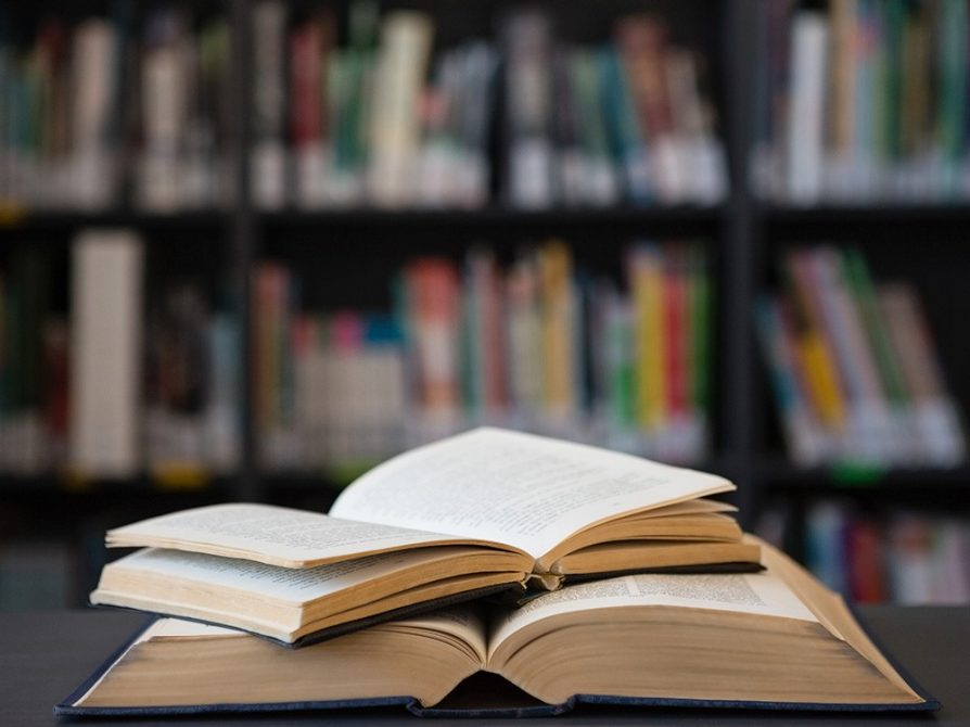 Two open books stacked on a table with bookshelves in the background.