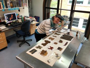 A woman with short, blond hair wearing a headband magnifier pushed up on her forehead leans over a metal table with several ancient, colorful textiles laid out flat. In the background is a desk with a computer, on which an image of a textile is displayed. 