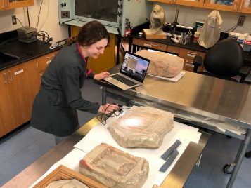 A woman with brown hair uses a handheld microscopy instrument attached to a computer. The tool shines a light onto a tan stone stela with carvings on its surface laid out on a table. Two other stelae with carvings are on the table. 