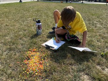 A middle school-aged child sits in the grass writing on a clipboard. Spread before them is a pile of fruit cereal.