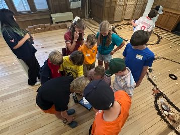 Kelsey staff member Chris Motz crouches on the ground with an iPad. Campers surround him to look at the screen.