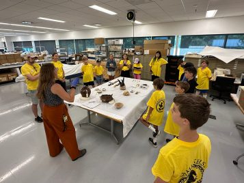 Fifteen middle schoolers and two chaperones stand around a large table covered in pots, bones, and other objects in UMMAA’s collections storage, listening to a woman speak.