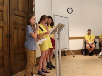 Three girls stand at the front of the room, holding sheets of paper. One child speaks into a microphone.