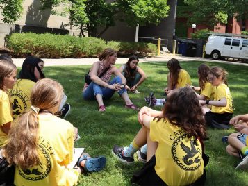 A group of 11 students and one tour guide sitting in a circle in the grass. Most of the kids wear bright-yellow shirts with the Camp Kelsey logo.