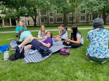 A group of middle school students and a camp counselor sit and sprawl on a picnic blanket and grass with their lunchboxes and drink bottles.
