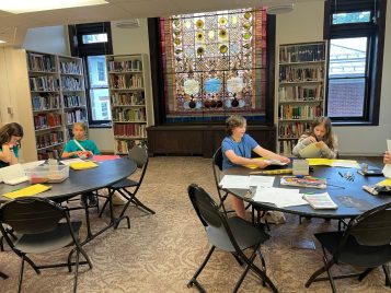 A small group of middle school students sit around two tables covered with craft supplies, folders, and paper in front of the stained-glass Tiffany window in the Kelsey Museum library. 