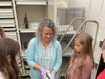 Wearing gloves, Michelle Fontenot holds out a small bundle containing a mummified bird to show a small group of middle school children.