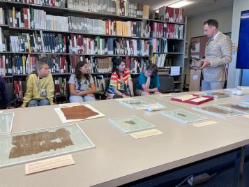 Brendan Haug holds a piece of papyrus up as he addresses campers. They sit in a row before a table covered with further papyrus examples encased in glass.