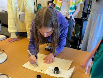 A middle schooler leans over a table to write on a sheet of papyrus using a nib pen and pot of ink.
