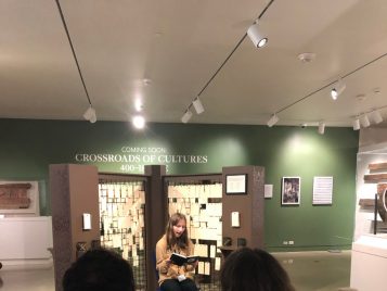 A middle school student sits in a chair in the Kelsey Museum’s special exhibition space reading aloud to a small audience.