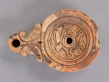 Buff clay oil lamp with a missing handle. Depictions of gladiatorial armor are carved into the surface: greaves, a helmet, a sword, and two shields.