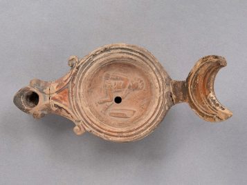 Ceramic oil lamp with a seated gladiator wearing a helmet and greaves, holding a sword. A curved shield is in front of him.