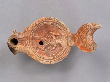 Pale buff clay oil lamp with reddish-brown glaze depicting a gladiator wearing a helmet, tunic, and greaves, with a shield on his arm. 