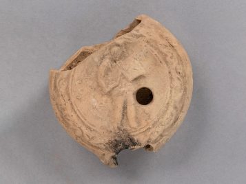 Buff clay oil lamp with its nozzle and handle broken off. A vine design closes an image of a gladiator standing, wearing a helmet and greaves and carrying a shield and dagger.