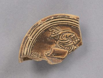 Fragment of a buff clay oil lamp with brown glaze showing the upper half of a gladiator wearing a helmet, tunic, and shield, carrying a curved sword. The front part of another gladiator is visible at one of the points of breakage.