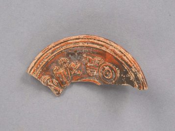 Fragment of a buff clay oil lamp with reddish-brown glaze. Visible around the discus is a circle of gladiatorial armor, including a greave, scutum, sica, helmet, breastplate, and shield.