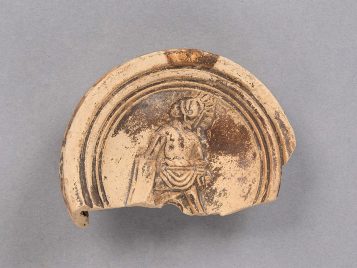 Fragment of a buff clay oil lamp with brown glaze. At the center of the object is a standing gladiator wearing a crested helmet, a loincloth, and an arm guard, holding a sword and long shield.