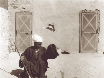 A man holding a stick sits on the ground, looking at two hooded snakes coiled before him.