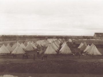 Rows of white tents making up a camp. Figures of refugees and donkeys can be seen in the foreground, while long, low buildings are visible at right.