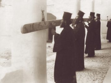Four monks wearing matching black attire stand in a row in a monastery courtyard, using mallets to strike a long, skinny piece of wood. A fifth monk stands alone in the back with a smaller bar of wood and a mallet.