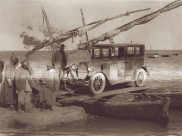 A 1920s sedan loaded atop a makeshift ferry consisting of fishing boats connected by ropes and planks. To the left stands a worker from the University of Michigan expedition and three Tunisian men.