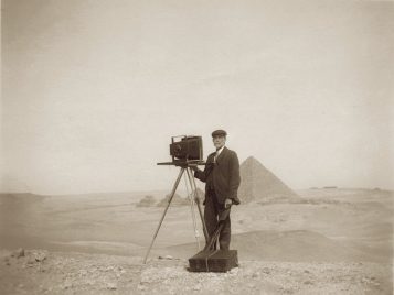 George Swain, dressed in a suit and hat and with a suitcase at his feet, stands with a large-format camera on a tripod in front of a row of four pyramids.