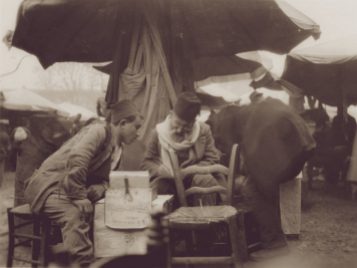 A young man wearing a fez leans forward to speak to an older man in a fez acting as a scribe in a market stall. 