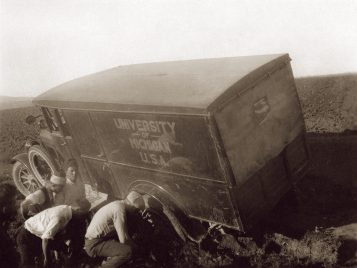 A 1924 Graham Brothers truck, printed with “University of Michigan, U.S.A.” on its side, leans at an angle with its lefthand wheels stuck in mud. A group of four men work to free the vehicle.