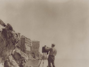George R. Swain faces stands among a rocky section of coastline, balancing his camera and tripod. His back is turned, capturing a photograph of a monastery and cliffside in the distance.