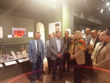 Janet Richards and Mohamed Ismail Khaled talk to one another in the Weni the Elder exhibit space, which displays a tall stone monument and a low vitrine full of objects. A crowd of people stands around them.