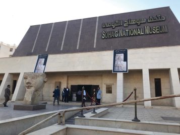 Exterior of the Sohag National Museum: a tan and brown building with “Sohag National Museum” written in English and Arabic, with a large stone head on a platform out in front. Banners advertising the Weni exhibit hang from the façade.