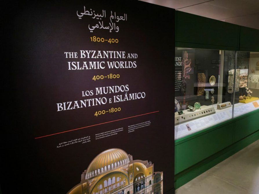 A black wall with white and yellow text reading “The Byzantine and Islamic Worlds: 400–1800” in Arabic, English, and Spanish above a cross-section of a mosque, near a long museum case with glass, ceramic, wood, and stone objects.