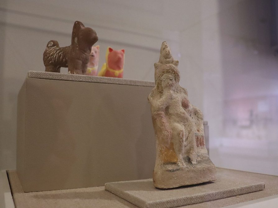 Four objects installed in the exhibition case: a ceramic figurine of Harpocrates with an amphora in his arm seated next to an oven, a dog with pointed ears and a curly tail, and two 3D replicas of the dog painted in red, pink, and yellow.