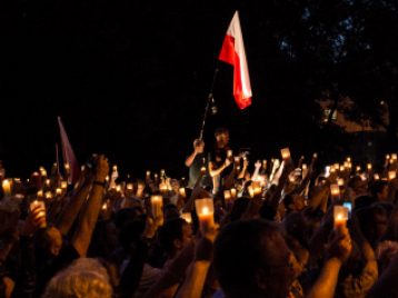 In this photograph taken at night, a man stands in the center of a crowd of people holding the red-and-white Polish flag. The people in the crowd are holding lit votive candles above their heads.