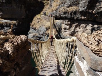 A plunging, long rope bridge crosses a cavern of rocks. A figure wearing a red hat is beginning to ascend the bridge, having walked across it.