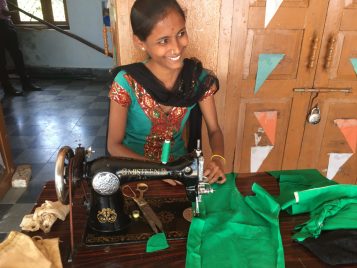A young woman wearing blue and red dress is sitting behind a sewing machine and smiling at someone outside the frame. Green cloth covers the table where shew orks.