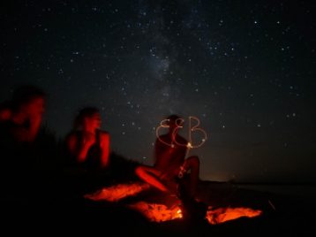 A dark photograph taken around a campfire. The red glow of the fire's embers is reflected against the people sitting around it.