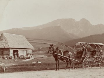 a black and white photo in a valley surrounded by mountains. There is a small building with a steep roof and a horse is harnessed to a buggy.