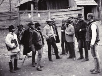 A photograph of a group of 12 men standing together talking. There is a tall wood fence behind them.