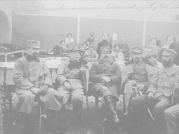 A photograph of a group of men sitting inside a large room. There is a row of six men in the front, wearing military uniforms.